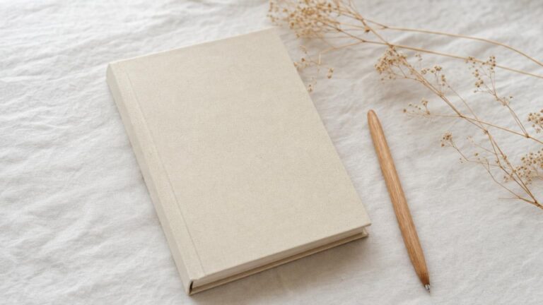 淡い色のリネンの上に置かれた、何も書かれていない開いたノートとペン、傍らの小さなドライフラワーのフラットレイ
