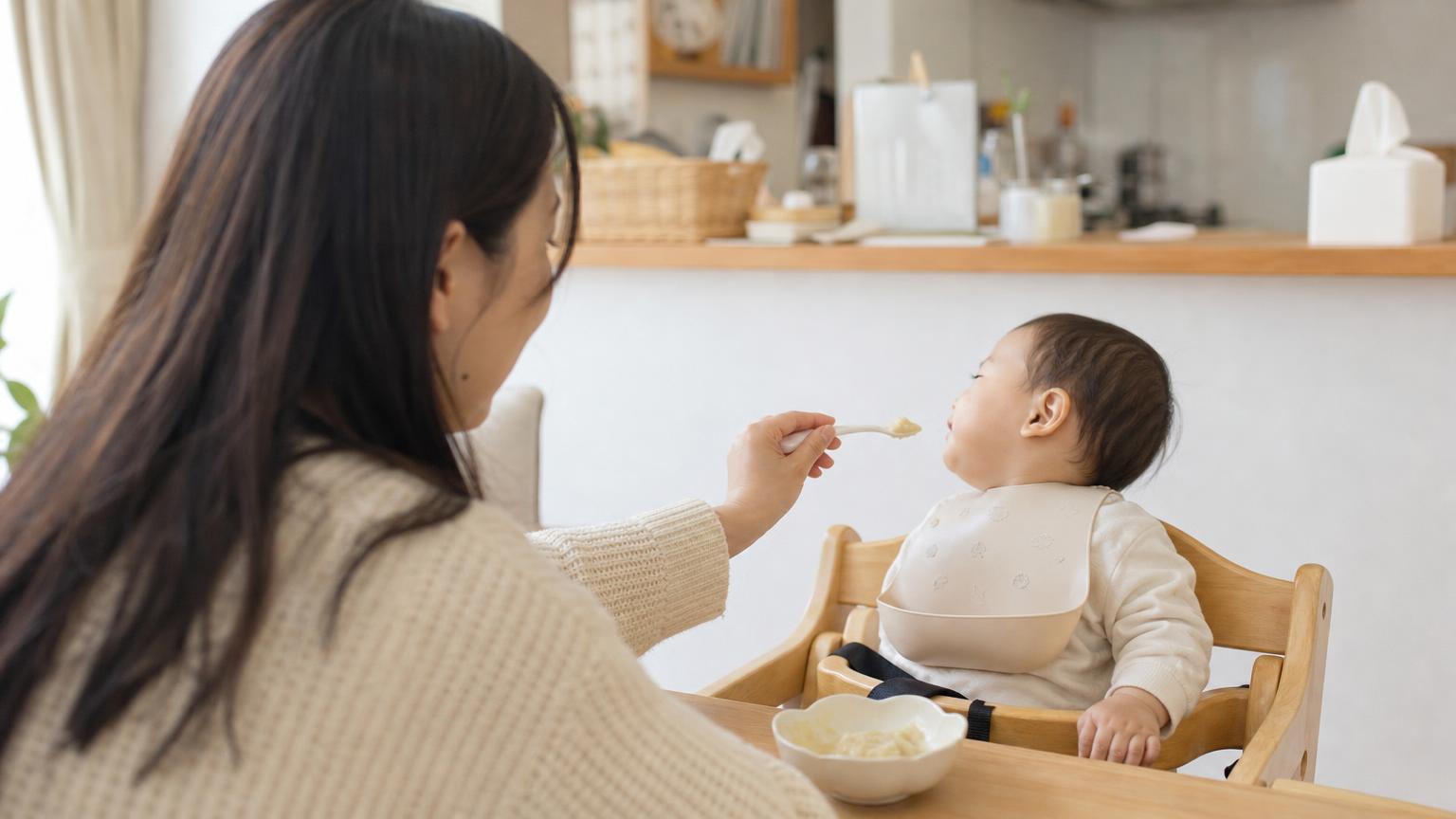 ハイチェアに座る赤ちゃんに、離乳食をスプーンで差し出す母親の手元。テーブルには小さな器が置かれている