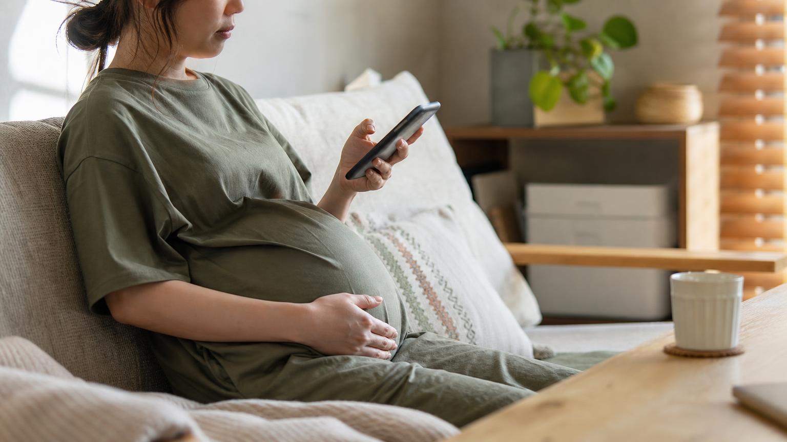 ソファに座り、片手でスマートフォンを持ち、もう片方の手をおなかにそえた妊婦。顔は写さず肩から下の構図で、午後のやわらかい光