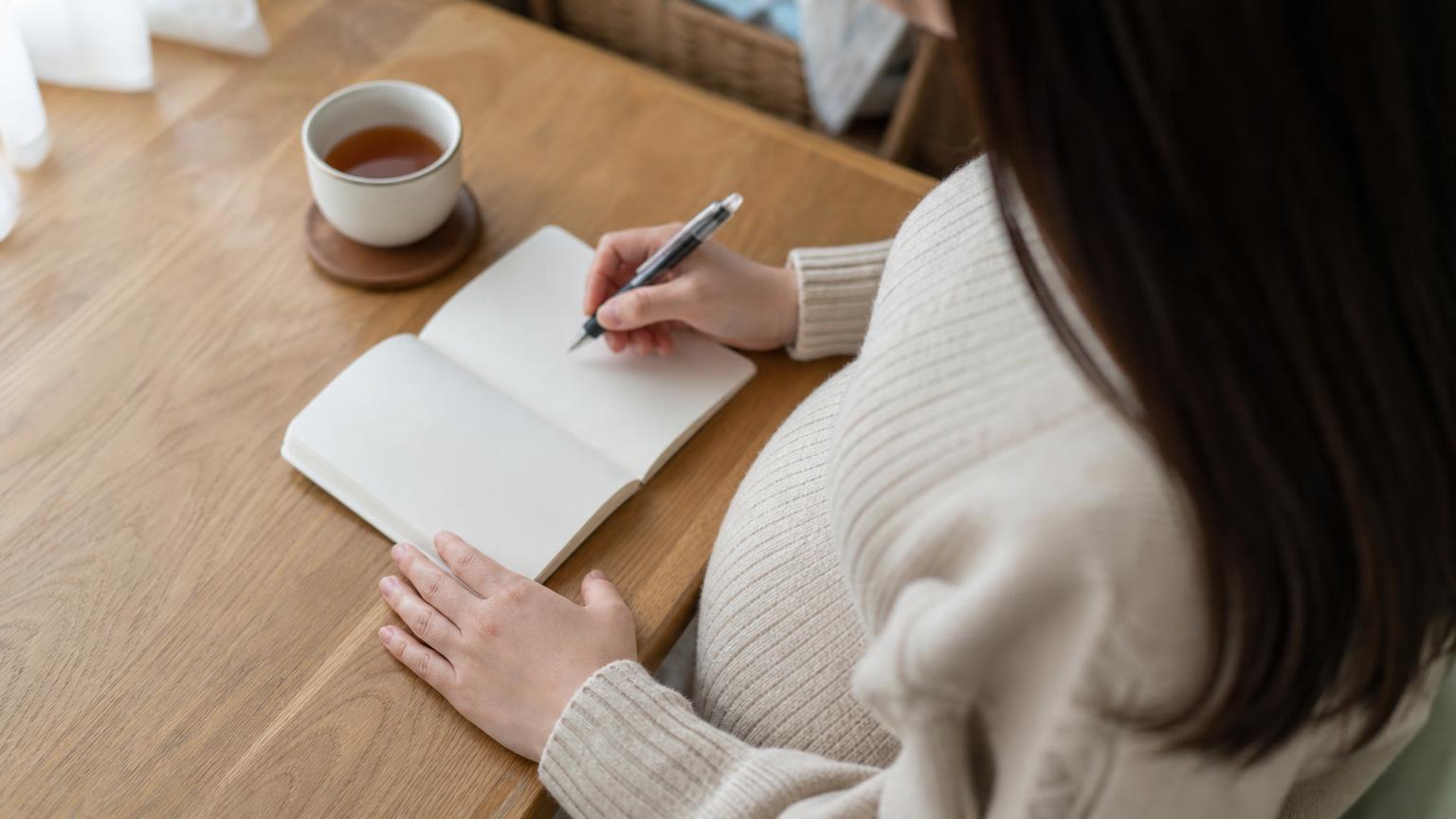 朝の台所のテーブルで小さなノートに体調を書き留める妊娠後期の女性の手元